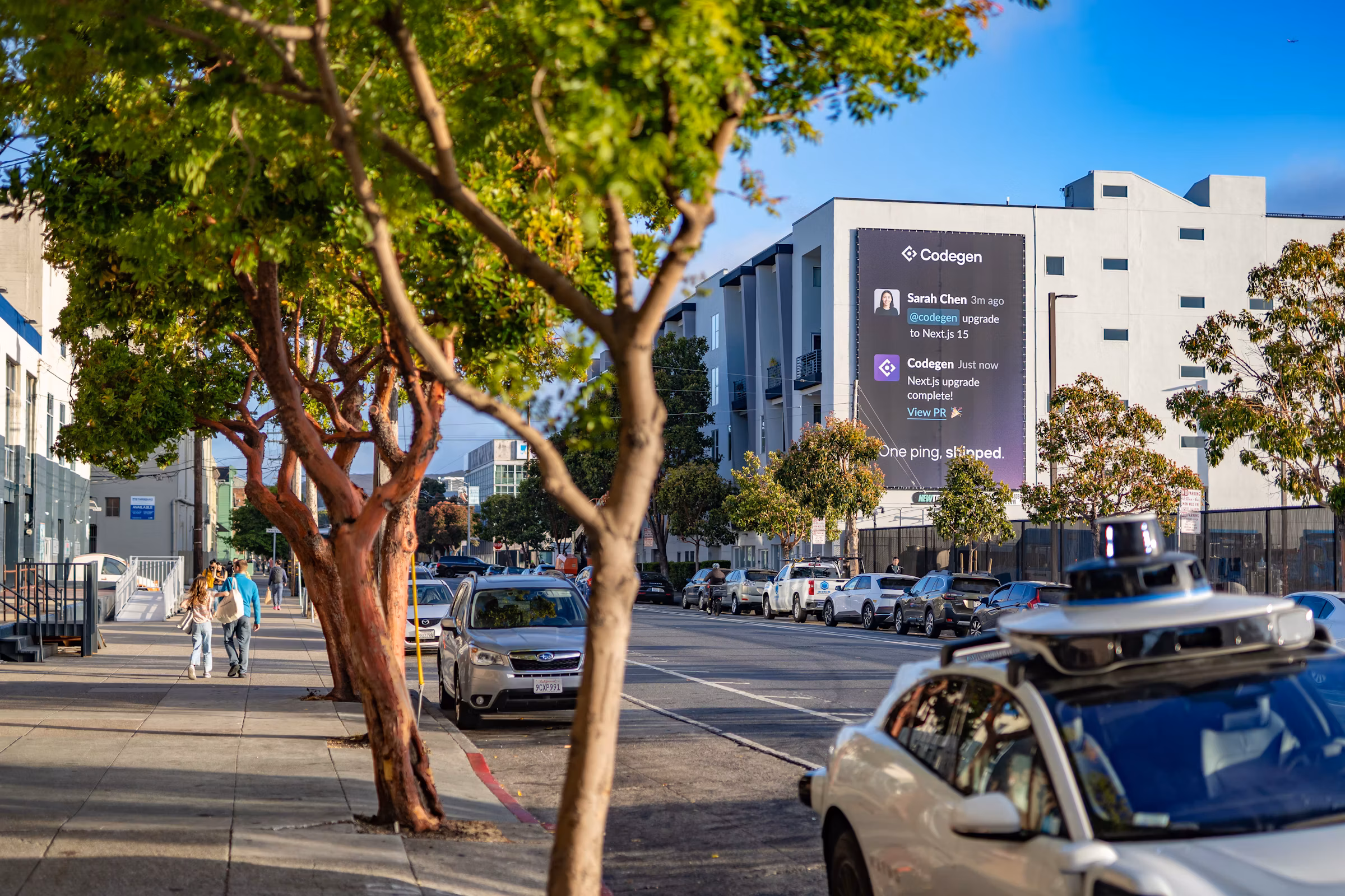 A building-side digital billboard showing a Codegen pull-request notification - 'One ping, shipped.' - as a sensor-topped self-driving car passes below at golden hour.